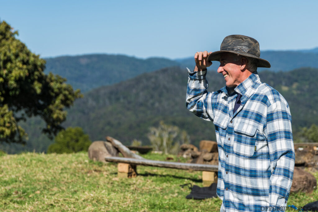 Photo of an adult male in a blue and white flannel shirt tipping his stetson with smile, presumable greeting someone off camera.