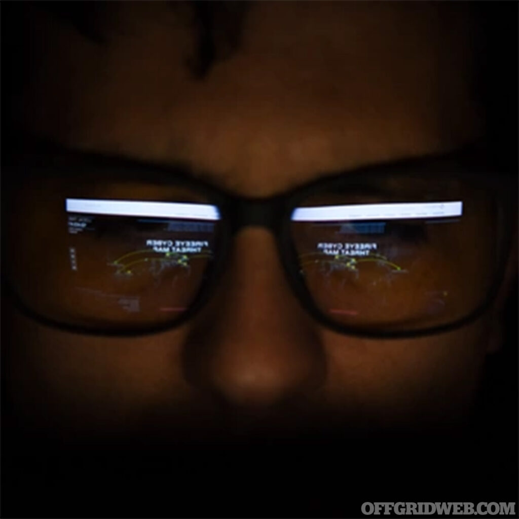 Photo of a nefarious cyber criminal checking the internet in a dark room, the glow of the computer reflecting in their glasses.