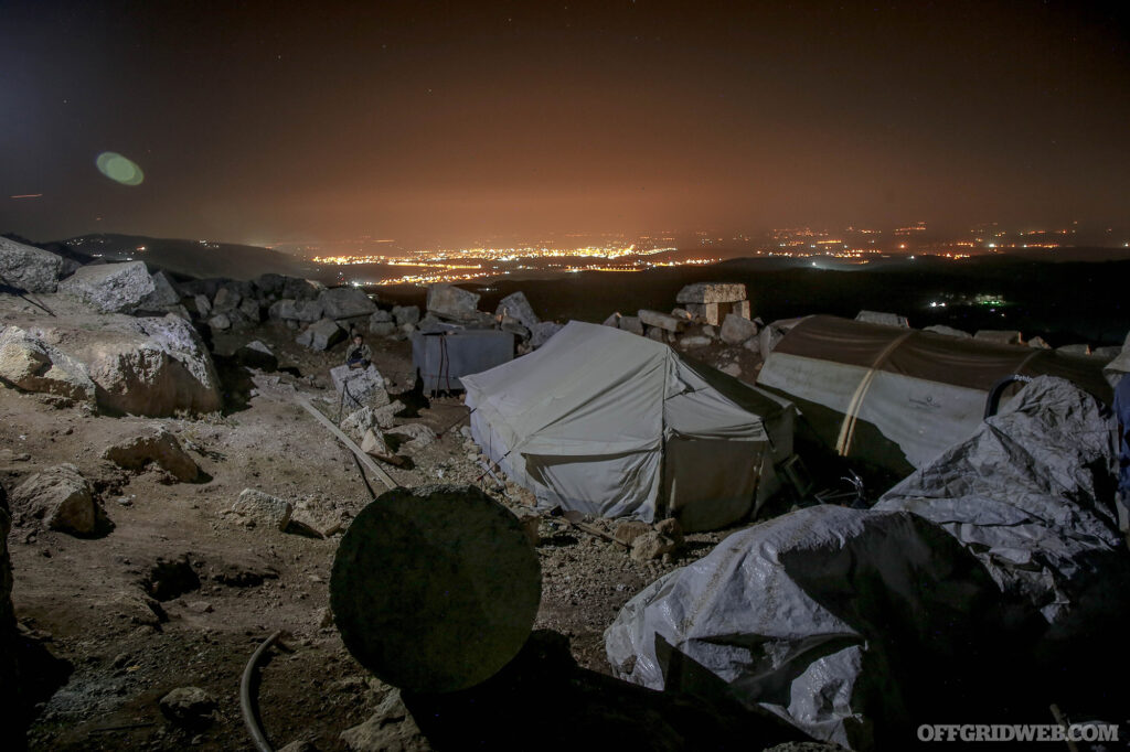 Photo of a refugee camp at night with city lights illuminating the sky in the background.