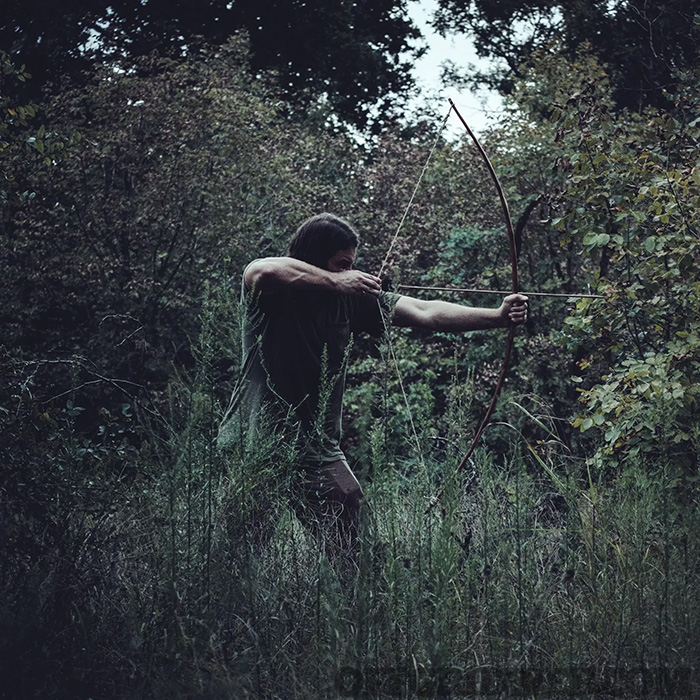 Photo of an adult male drawing back a long bow in the middle of a forest.
