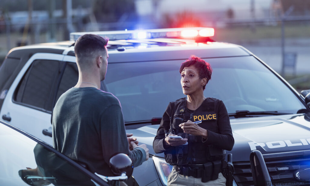 A policewoman taking a statement from a civilian outside her patrol car. The officer is a mature African-American woman in her 40s. She is talking with a young man in his 20s.