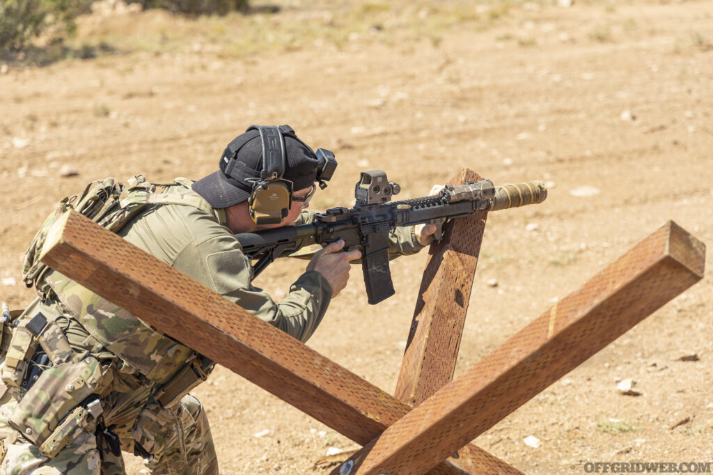 An adult male shooting from a crouched position behind an obstacle.