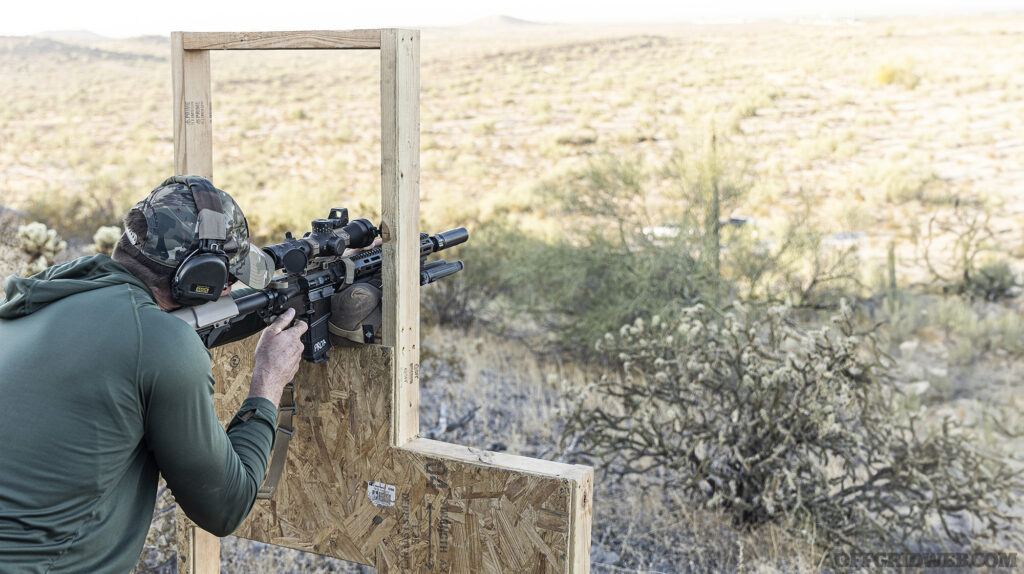 Photo of a man braced in a window mock up taking aim through a scoped rifle.