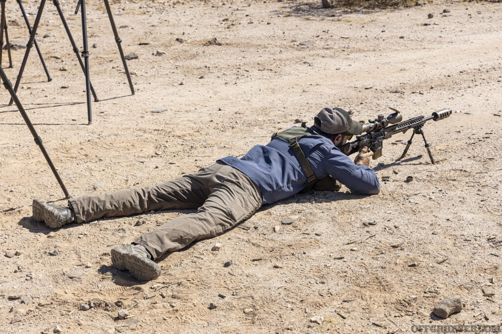 Photo of man lying in the prone on the dusty desert ground taking aim behind his rifle.