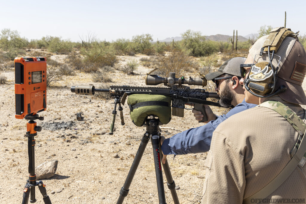 A marksman takes aim through a scoped carbine mounted on a tripod.