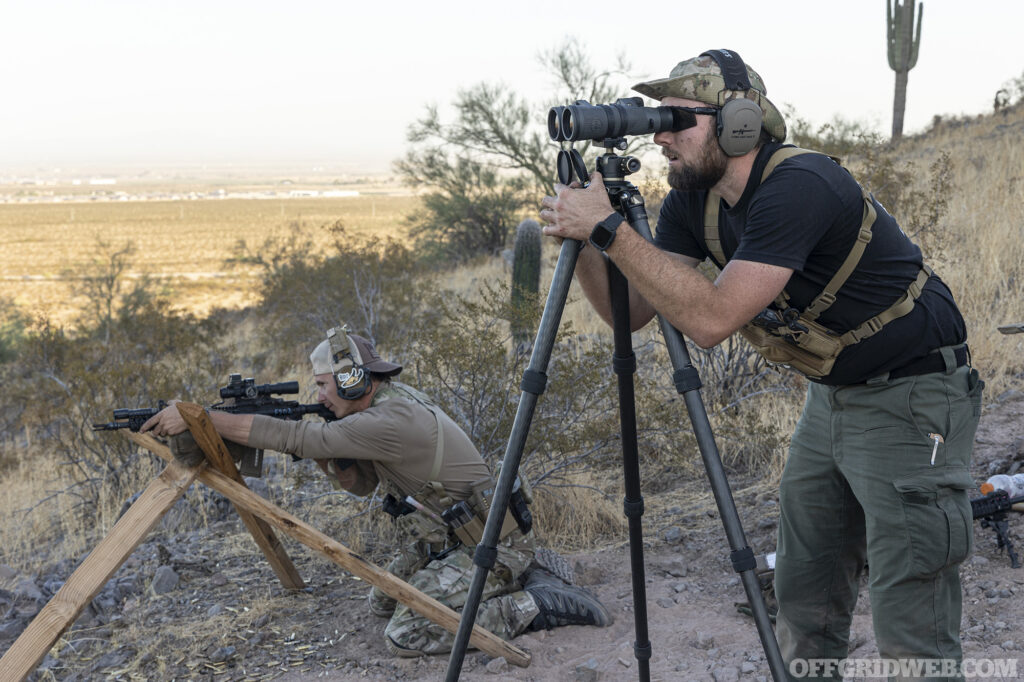 A man standing behind a spotting scope mounted to a tripod.