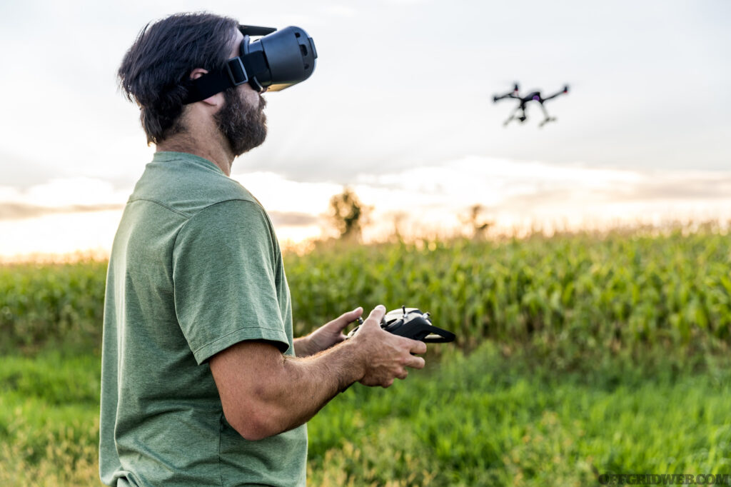 DSLR picture of a man flying a drone over a field by a beautiful summer sunset. The man is wearing Virtual Reality Goggles Headset.