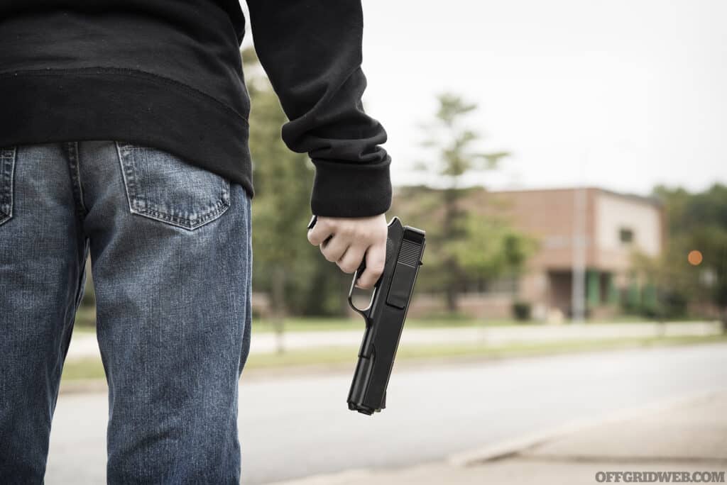 A student wearing jeans and a black hoodie stands outside of school holding a gun.